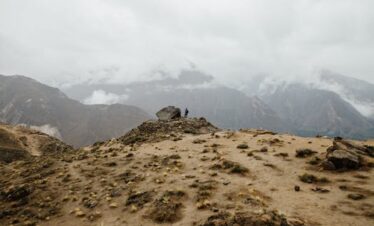 A lone figure standing on a rugged mountain amidst misty clouds at dawn.