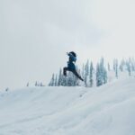 A man leaps across a snow-covered hillside against a backdrop of trees in Gulmarg.